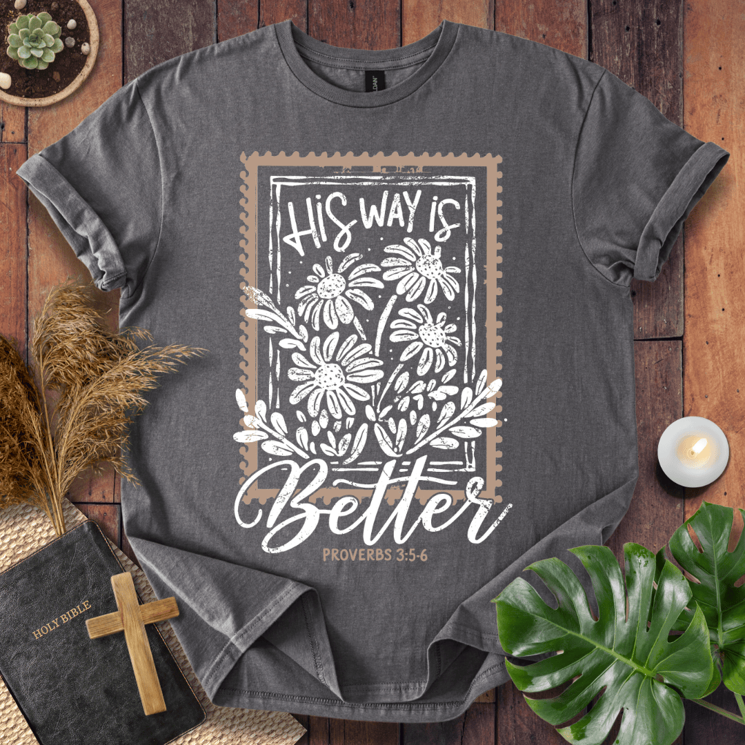 A dark gray graphic t-shirt laid flat on a rustic wooden surface, surrounded by decorative props including a Bible, a cross, dried grass, a small candle, and green leaves. The t-shirt features a white floral design with a brown border and the text 'HIS WAY is Better PROVERBS 3:5-6'.