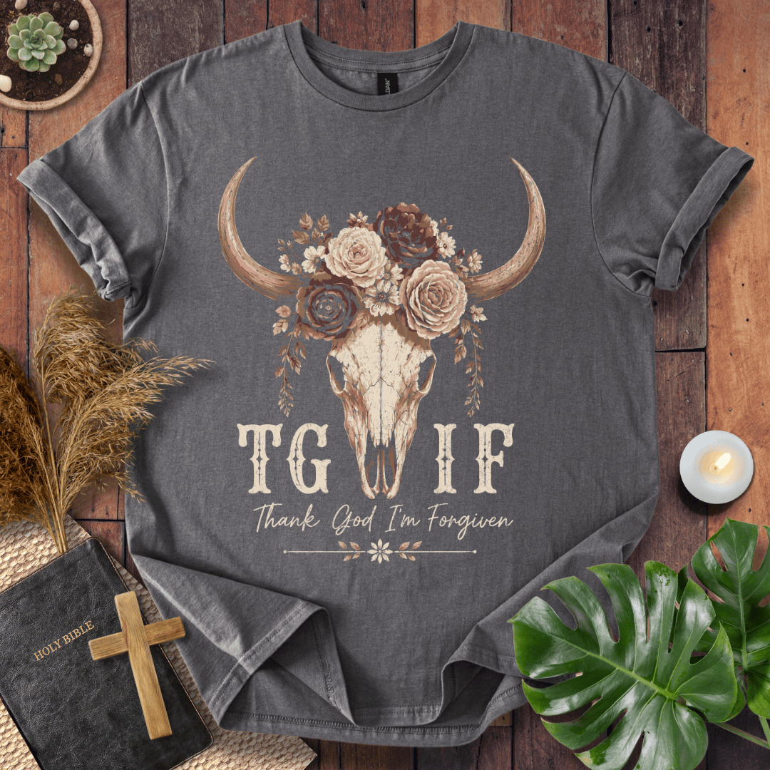 A dark gray graphic t-shirt laid flat on a rustic wooden background, surrounded by decorative props including a Bible with a wooden cross, dried grass, a small candle, and a green monstera leaf. The t-shirt features a central graphic of a skull with large horns decorated with flowers, and the text 'TG IF' and 'Thank God I'm Forgiven'.