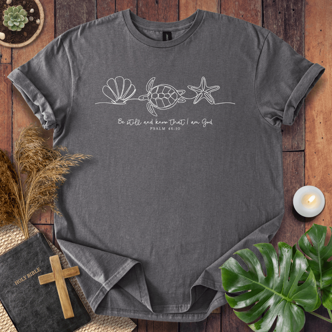 A dark gray crewneck t-shirt with a white line art graphic of a sea turtle, starfish, and seashell, with the text 'Be still and know that I am God' from Psalm 86:10. The shirt is displayed in a flat-lay arrangement on a rustic wooden background with a Bible, cross, pampas grass, a small candle, and a green monstera leaf.