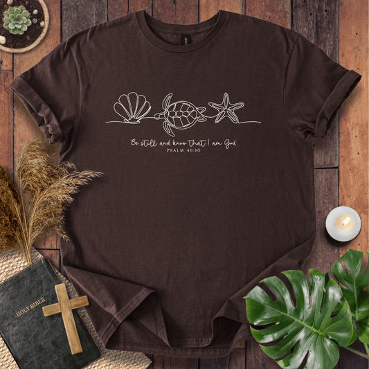 A dark brown short-sleeve t-shirt with a white line art graphic of a sea turtle, seashell, and starfish, with the Bible verse 'Be still and know that I am God Psalm 46:10' printed on the chest. The shirt is displayed in a styled flat-lay on a wooden background with a book, cross, dried grass, a candle, and a green plant leaf.