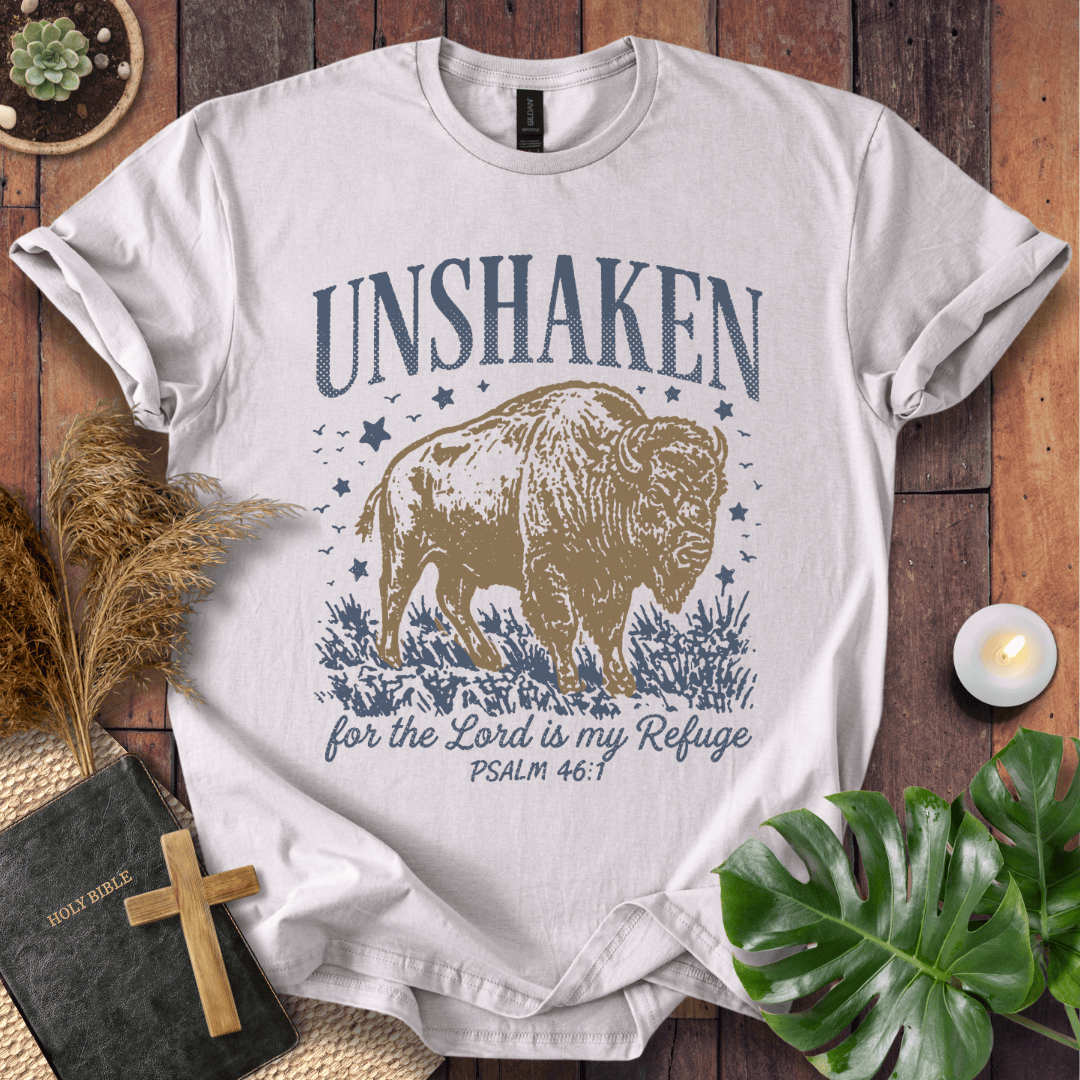 A white graphic t-shirt featuring a bison and the text 'UNSHAKEN for the Lord is my Refuge PSALM 46:1' in navy blue and tan, styled in a flat-lay on a wooden background with a bible, cross, pampas grass, and green leaves.