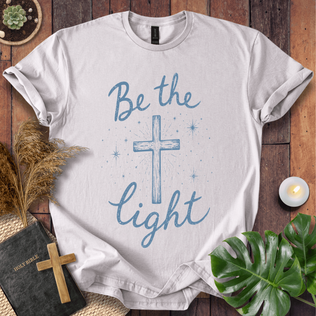 Christian t shirt for women with blue "Be the Light" text, a central cross, and starry accents on a wooden background.