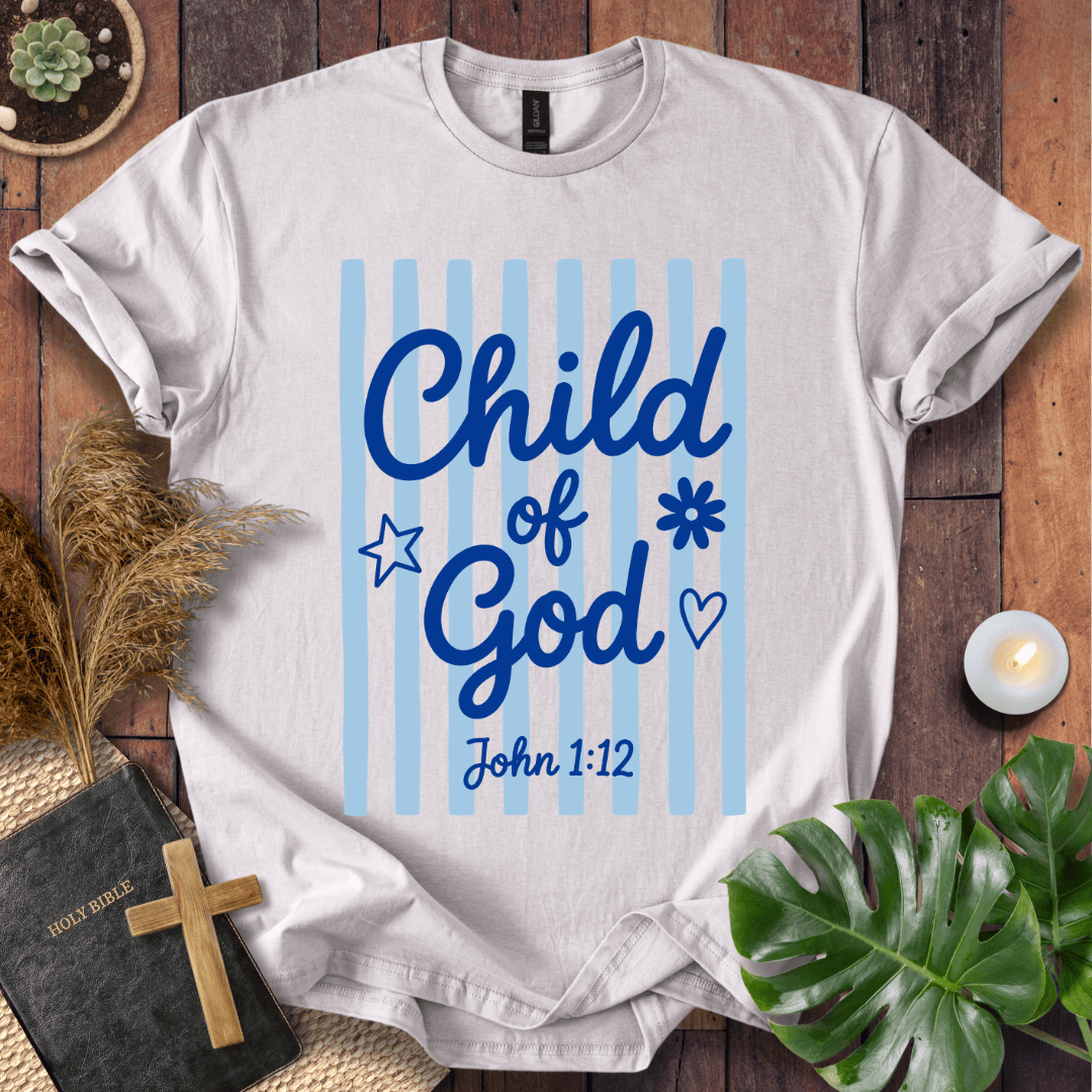 A white short-sleeve T-shirt laid flat on a rustic wooden surface, featuring a blue graphic that reads 'Child of God' with 'John 1:12' below it, alongside light blue vertical stripes and small decorative icons. The shirt is styled with a Bible, cross, pampas grass, candle, and green leaves around it.