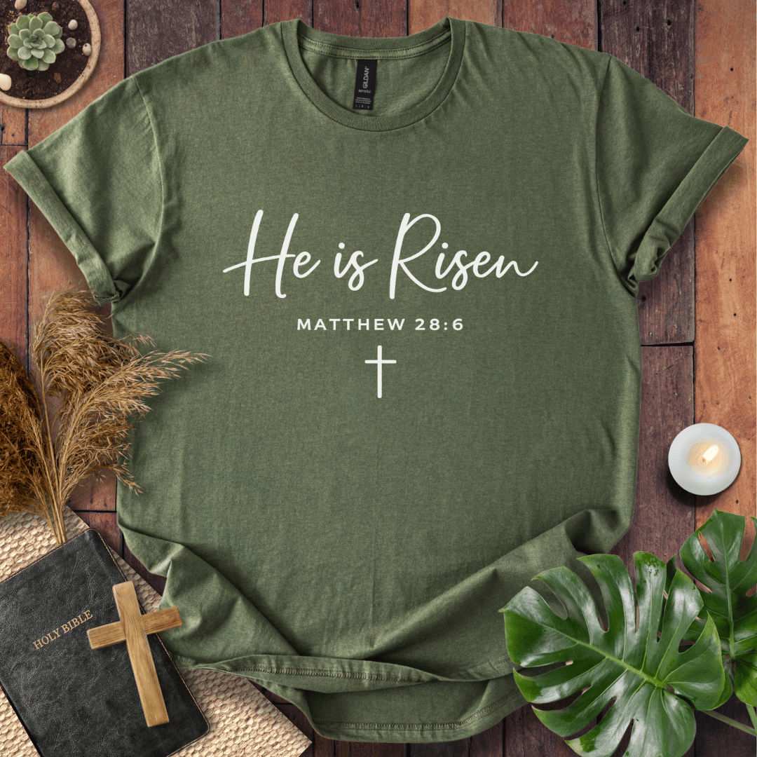 Easter Christian t-shirt with white "He is Risen" script and cross, styled on wood with a Bible and wooden cross.