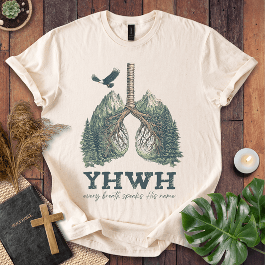 Christian t-shirt with YHWH design, tree-lung illustration, mountains, and “Every Breath Speaks His Name” text, laid flat with Bible and plants.