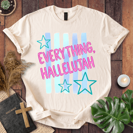 A cream-colored graphic t-shirt laid flat on a dark wood background, surrounded by decorative props including a Bible, a wooden cross, pampas grass, a candle, and green leaves. The t-shirt features the text 'EVERYTHING, HALLELUJAH' in pink bubble letters over light blue and cyan vertical stripes, accented with two outlined stars.