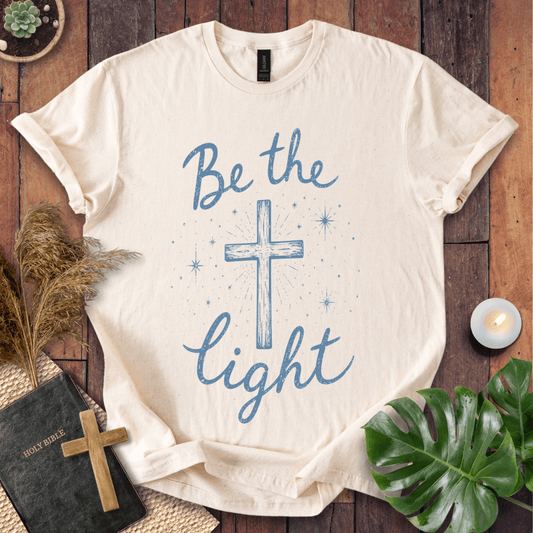 Christian t shirt for women with blue "Be the Light" text, a central cross, and starry accents on a wooden background.