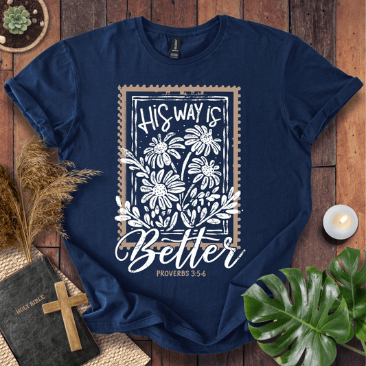 A navy blue graphic t-shirt laid flat on a rustic wooden surface, surrounded by decorative props including a Bible with a wooden cross, dried grass, a small candle, and green leaves. The t-shirt features a white floral stamp-style graphic with the text 'HIS WAY IS Better PROVERBS 3:5-6'.