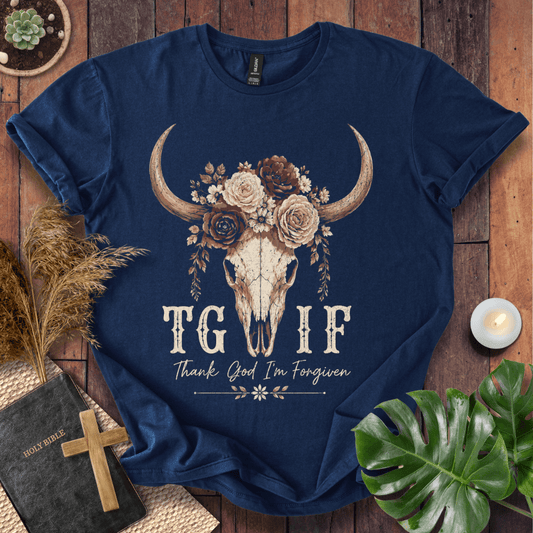 A navy blue graphic t-shirt laid flat on a rustic wooden background with decorative props including a book, cross, dried grass, monstera leaf, and candle. The shirt features a large floral and horned skull graphic above the text 'TG IF' and 'Thank God I'm Forgiven'.