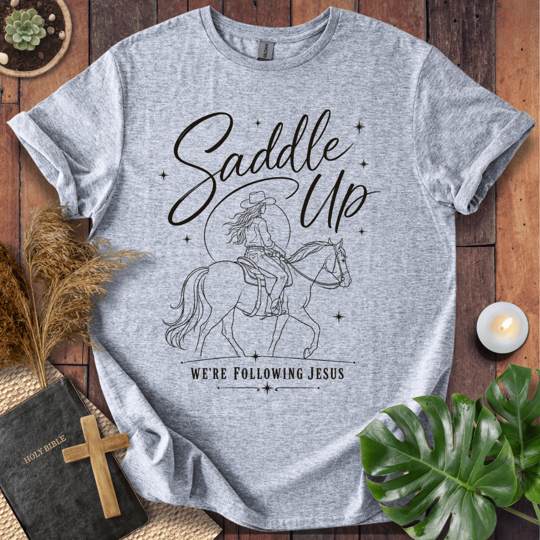 "Saddle Up, We're Following Jesus" shirt with a cowgirl riding a horse under a moonlit sky, displayed on a wooden background.