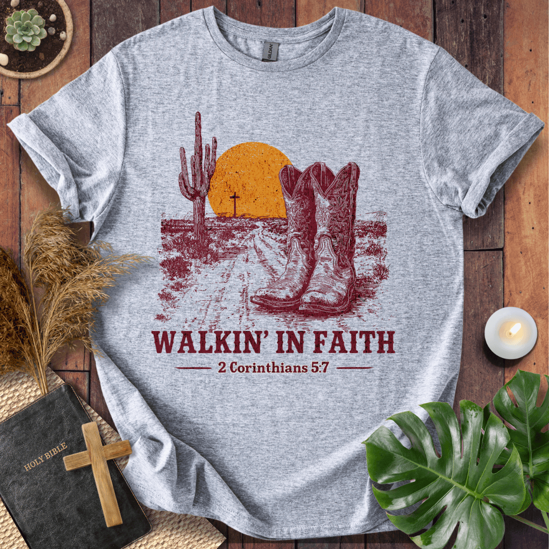 Gray t-shirt with cowboy boots and cactus design on a wooden surface with plants and a Bible.
