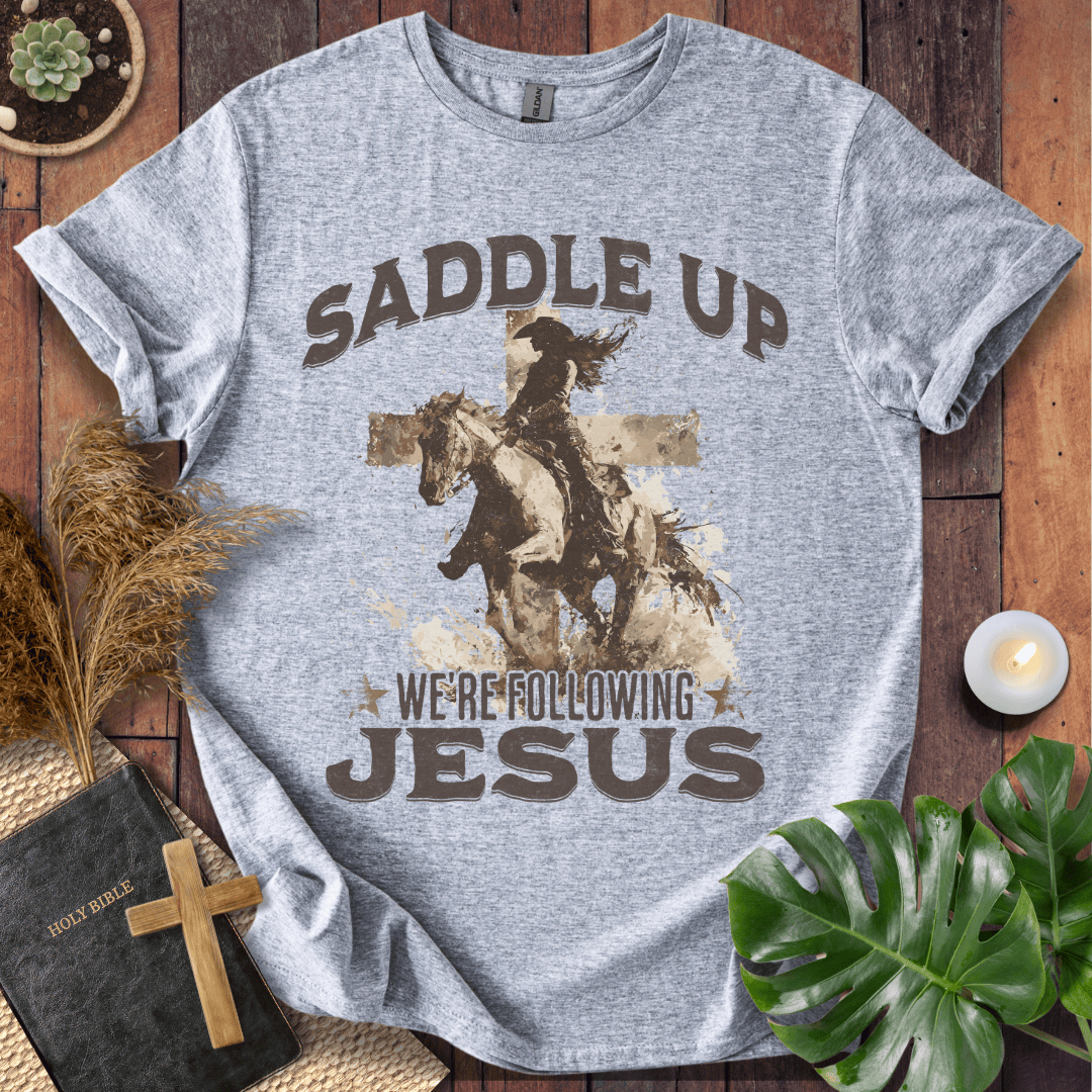 t-shirt featuring a cowgirl riding a horse in front of a cross with the text Saddle Up We're Following Jesus.