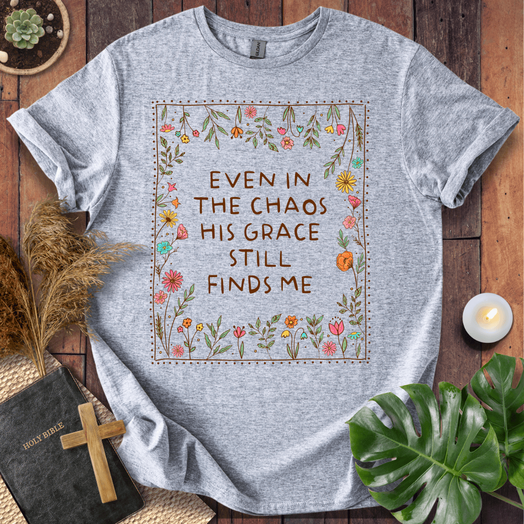 A heather gray graphic t-shirt laid flat on a rustic wooden surface, surrounded by decorative props including a Bible with a wooden cross, a lit candle, dried grass, and green leaves. The t-shirt features a central graphic with the text 'EVEN IN THE CHAOS HIS GRACE STILL FINDS ME' surrounded by a border of colorful flowers and green leaves.