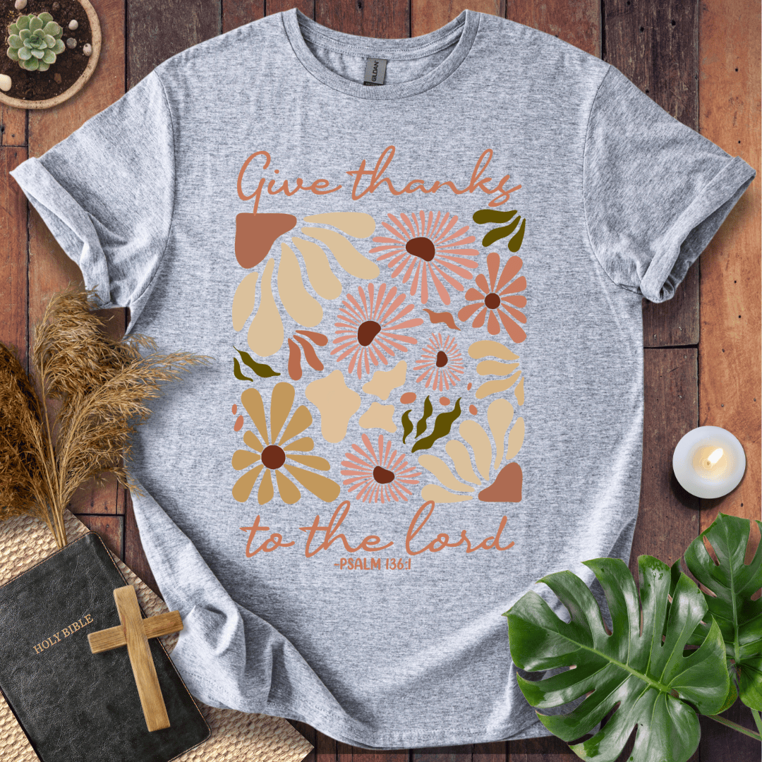 Give Thanks to the Lord Psalm 136:1 white graphic t-shirt with retro boho rust and sage wildflowers on a wood background.