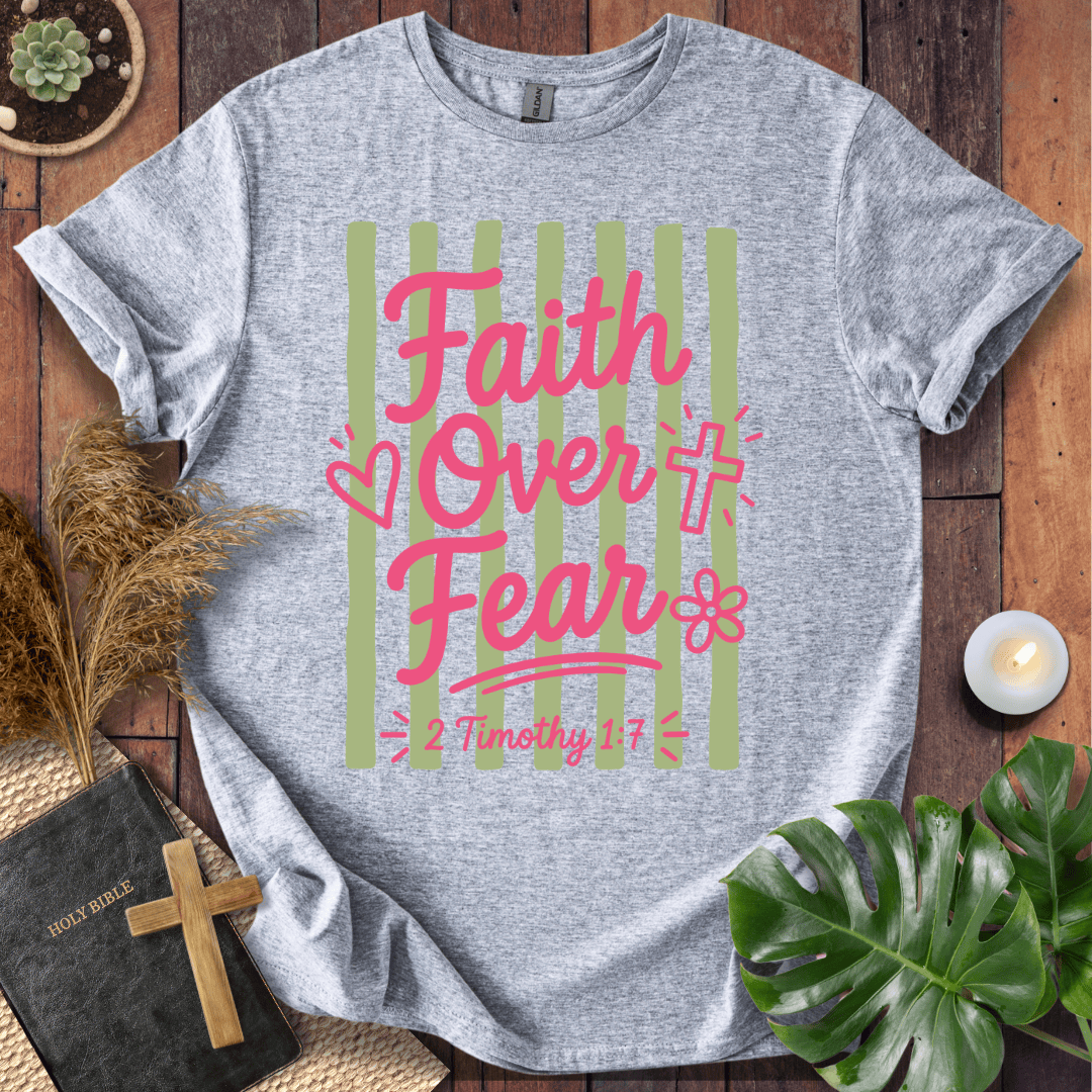 A heather gray graphic t-shirt laid flat on a rustic wooden surface, surrounded by decorative props including a Bible, cross, pampas grass, a lit candle, and a green monstera leaf. The shirt features a pink and light green striped graphic with the text 'Faith Over Fear' and '2 Timothy 1:7'.