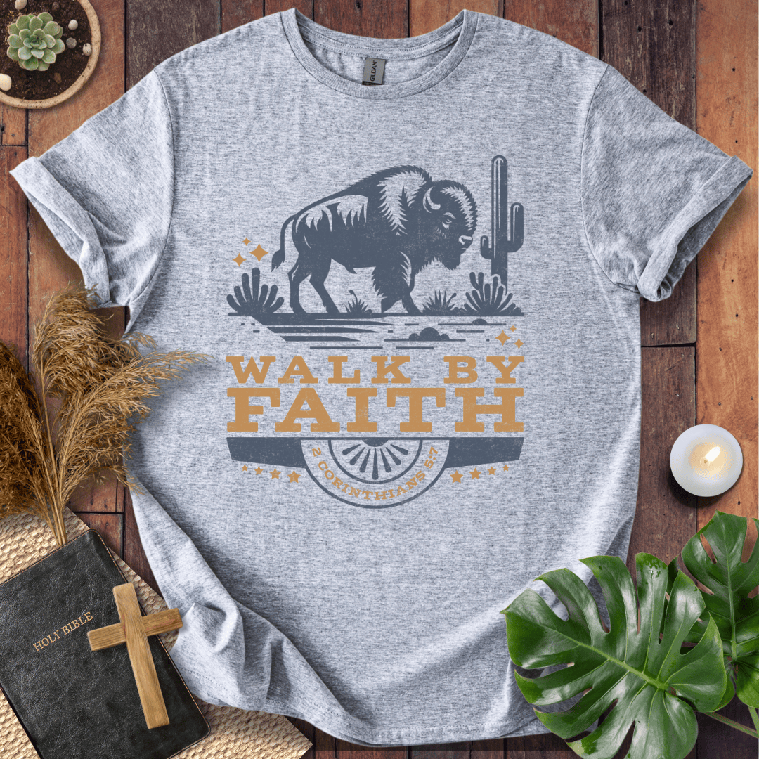 Gray t-shirt with a graphic of a bison and cactus, and 'Walk by Faith' text, on a wooden surface with decorative items.