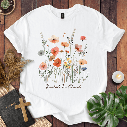 A white short-sleeve T-shirt laid flat on a rustic wooden surface, featuring a watercolor-style floral print in soft pink, orange, yellow, blue, and green tones above the words 'Rooted In Christ' in dark script. Decorative props including a Bible with a wooden cross, dried grass, a lit candle, and green leaves surround the shirt.