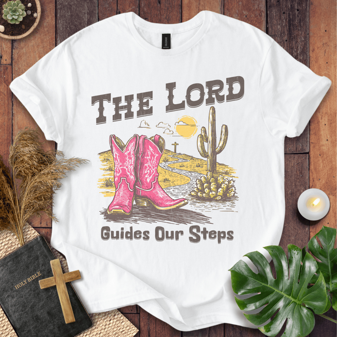 t-shirt with pink cowboy boots and "The Lord Guides Our Steps" text in a desert landscape with a cross and cactus.
