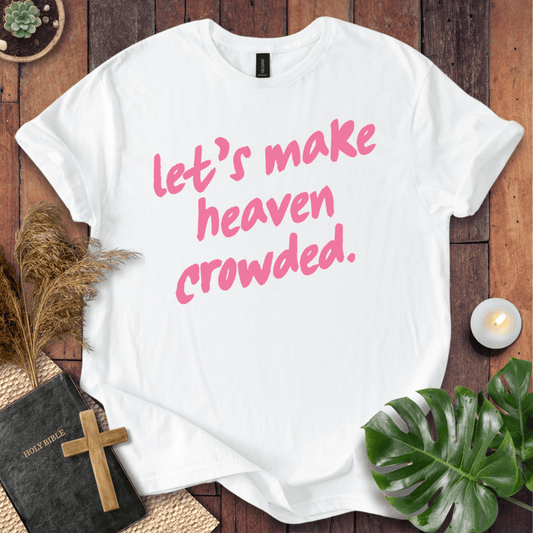 A white short-sleeve T-shirt laid flat on a rustic wooden surface, printed with the phrase "let's make heaven crowded." in pink script.