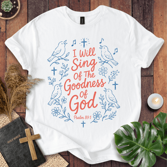 A white short-sleeve T-shirt laid flat on a rustic wooden surface, featuring blue line-art birds, flowers, and crosses with the phrase 'I Will Sing Of The Goodness Of God' and 'Psalm 89:1' in coral-red lettering. Decorative props including a book with a cross, dried grass, a candle, and green leaves surround the shirt.