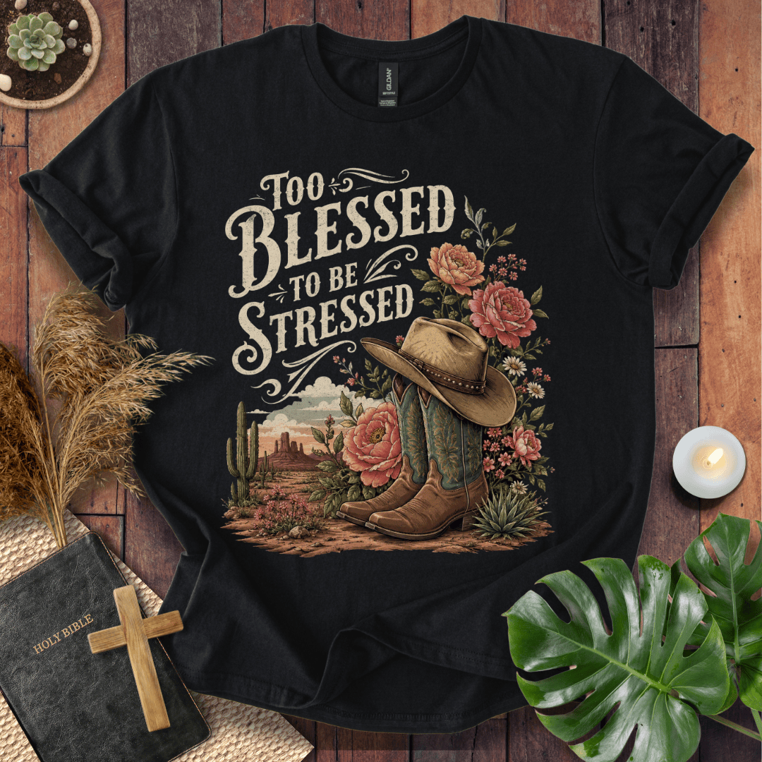 A black short-sleeve graphic T-shirt laid flat on a rustic wood surface, surrounded by decorative props including a Bible with a wooden cross, dried grass, a candle, and green leaves. The shirt features a large western-themed print with the phrase 'Too Blessed to Be Stressed,' flowers, boots, a cowboy hat, cactus, and a desert landscape.