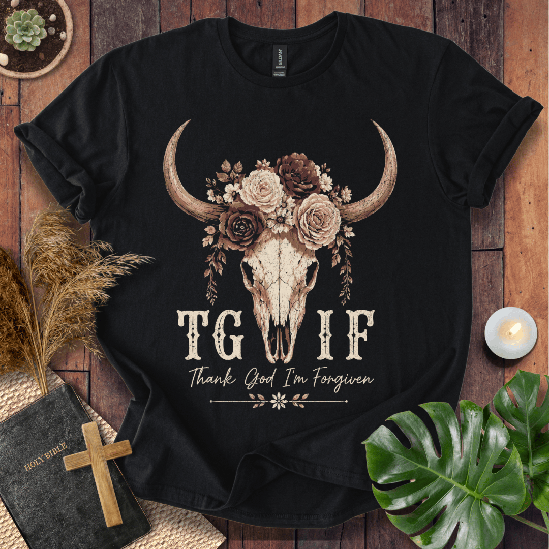 A black short-sleeve graphic T-shirt laid flat on a rustic wood surface, surrounded by decorative props including a Bible with a wooden cross, dried grass, a small candle, and green leaves. The shirt features a large central graphic of a skull with long curved horns, decorated with roses and flowers, above the text 'TGIF' and 'Thank God I'm Forgiven'.