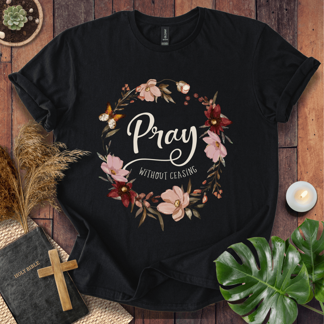 "Pray Without Ceasing" text inside a floral wreath with a butterfly on a shirt next to a Bible and cross.