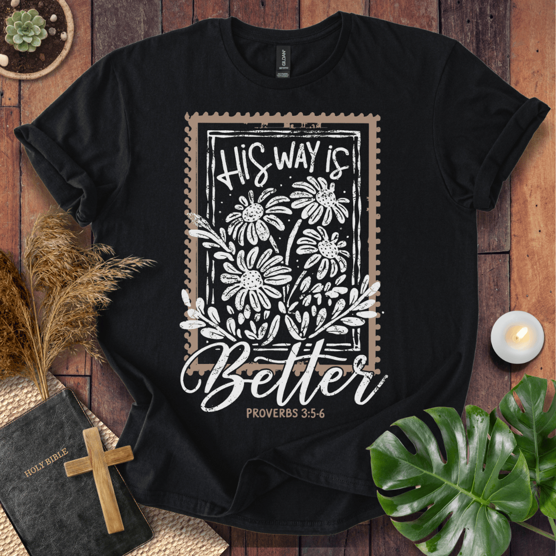 A black short-sleeve graphic T-shirt laid flat on a rustic wood surface, surrounded by decorative props including a Bible, cross, dried grass, candle, monstera leaf, and small plant. The shirt features a large rectangular floral print with the words “His Way Is Better” and “Proverbs 3:5-6” in white script and line art.