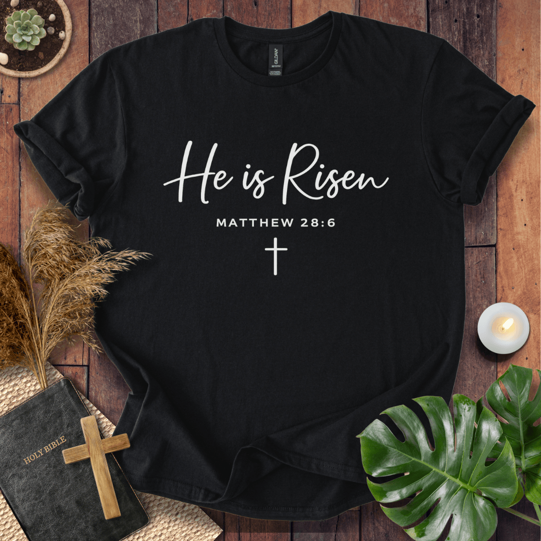 Easter Christian t-shirt with white "He is Risen" script and cross, styled on wood with a Bible and wooden cross.