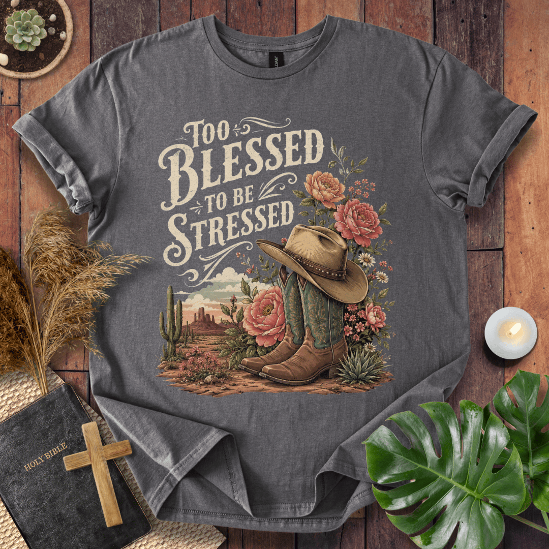 A dark gray graphic t-shirt laid flat on a rustic wooden background, surrounded by decorative props including a Bible with a cross, a candle, dried grass, and green leaves. The t-shirt features a western-themed graphic with the text 'Too Blessed to be Stressed' and an illustration of cowboy boots, a cowboy hat, cacti, and pink flowers.