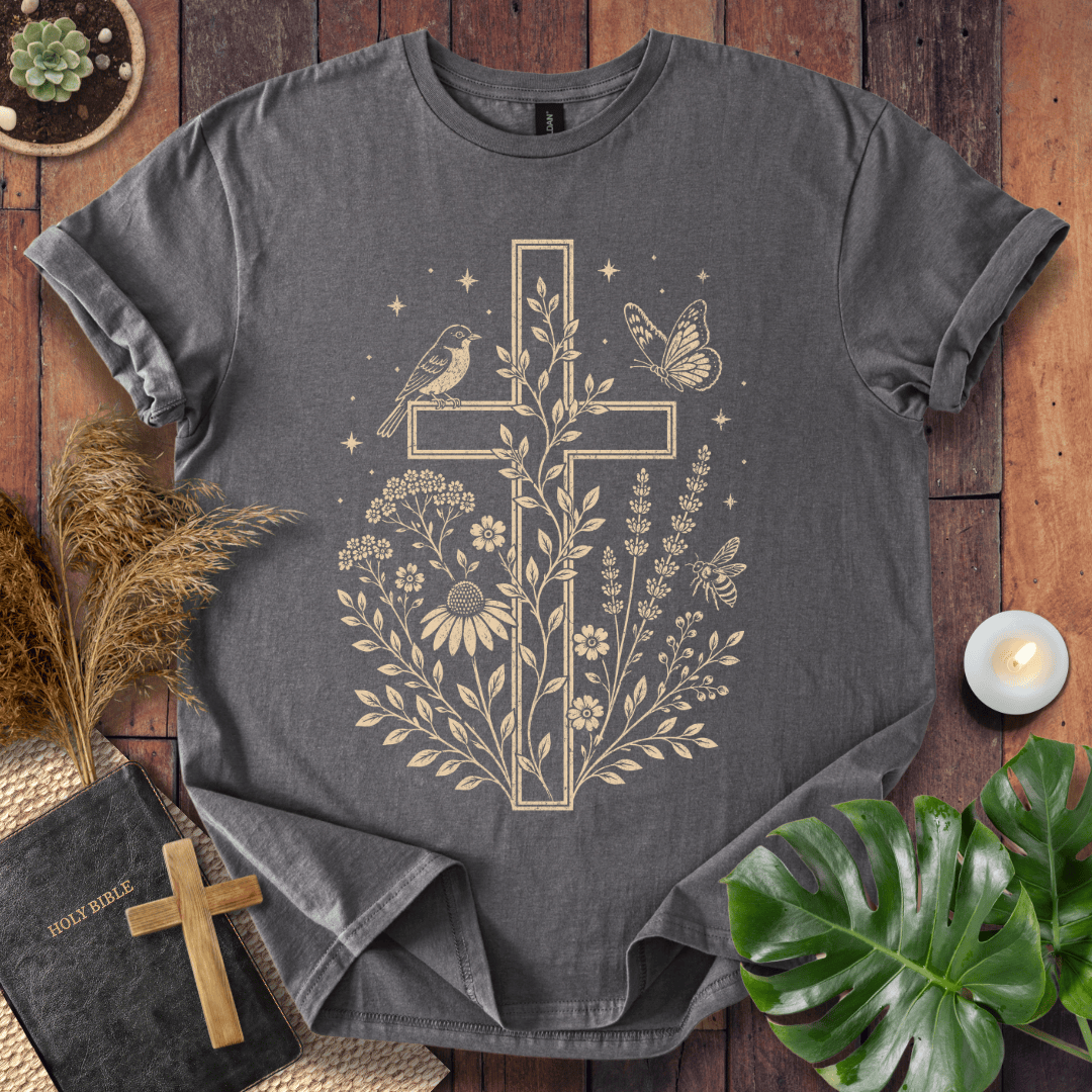 A dark gray t-shirt with a beige graphic of a cross decorated with flowers, leaves, a bird, and a butterfly, laid flat on a rustic wooden background with decorative props including a Bible, a candle, and green leaves.