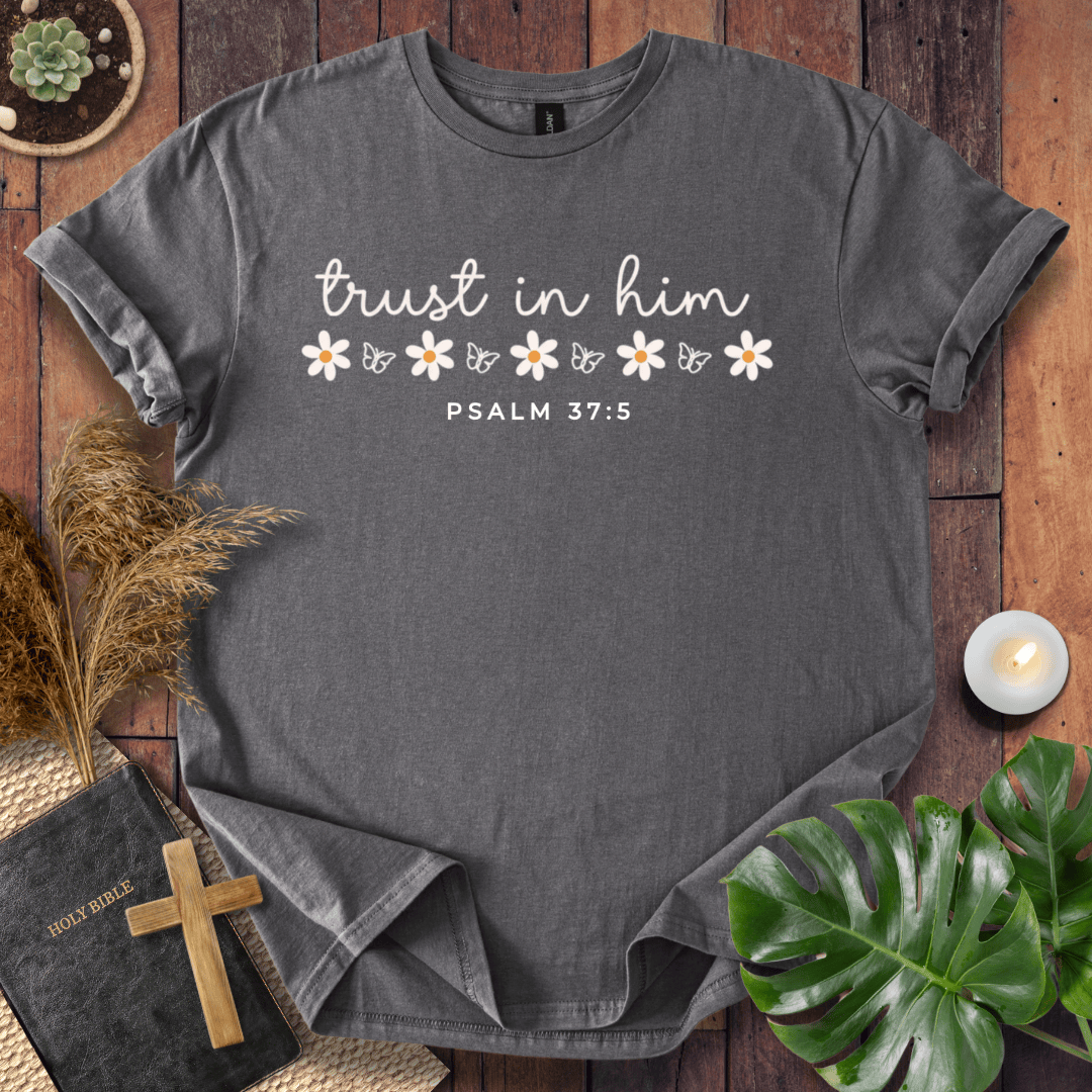 A dark gray heather crewneck t-shirt with 'trust in him' and 'PSALM 37:5' printed in white, decorated with white daisies and butterflies. The shirt is styled in a flat-lay arrangement on a wooden surface with a bible, cross, pampas grass, a small plant, and a lit candle.