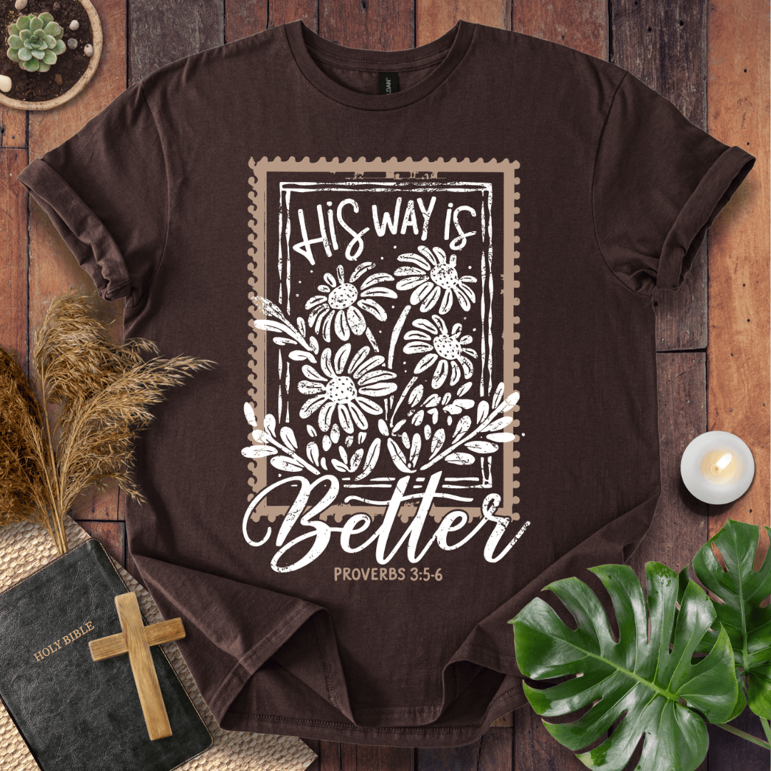 A dark brown short-sleeve graphic T-shirt laid flat on a rustic wood surface, surrounded by decorative props including a Bible with a wooden cross, dried grass, a small candle, green leaves, and a succulent. The shirt features a large white and beige stamp-style floral graphic with the text 'His Way Is Better' and 'Proverbs 3:5-6'.