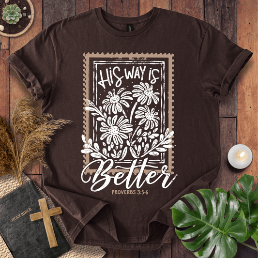 A dark brown short-sleeve graphic T-shirt laid flat on a rustic wood surface, surrounded by decorative props including a Bible with a wooden cross, dried grass, a small candle, green leaves, and a succulent. The shirt features a large white and beige stamp-style floral graphic with the text 'His Way Is Better' and 'Proverbs 3:5-6'.