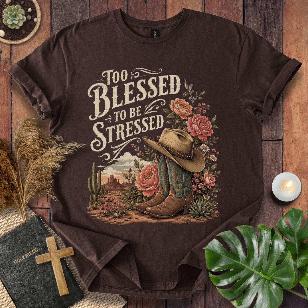 A dark brown graphic t-shirt laid flat on a rustic wooden background, surrounded by decorative props including a Bible with a wooden cross, a lit candle, dried grass, and a green monstera leaf. The t-shirt features a western-themed graphic with the text 'Too Blessed to be Stressed' in a distressed cream font, accompanied by a cowboy hat, boots, cactus, and pink roses.