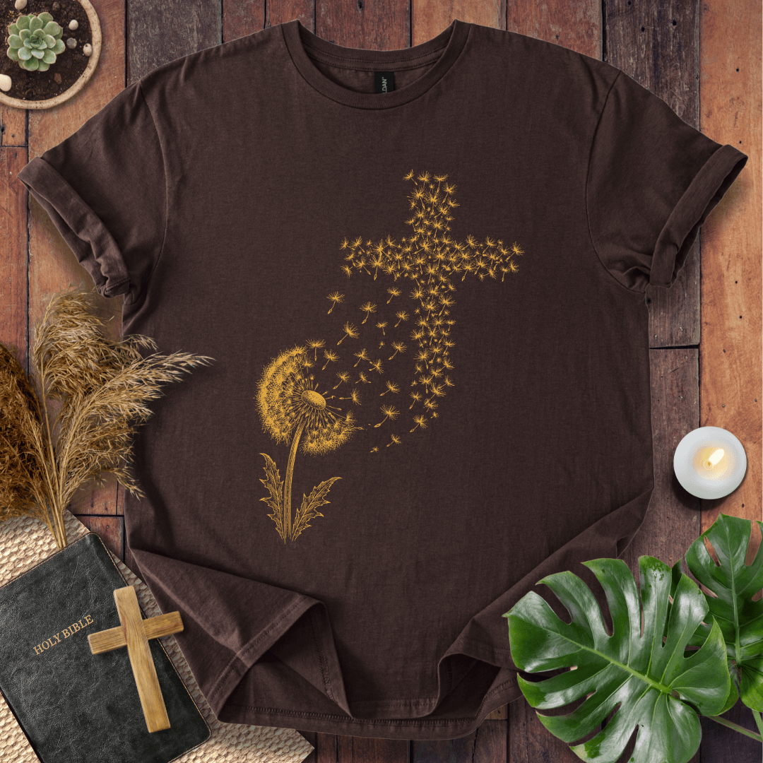 graphic t-shirt featuring a golden dandelion with seeds blowing in the wind to form a Christian cross shape.
