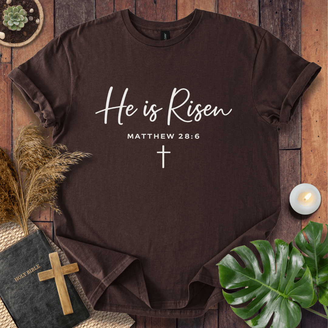 Easter Christian t-shirt with white "He is Risen" script and cross, styled on wood with a Bible and wooden cross.
