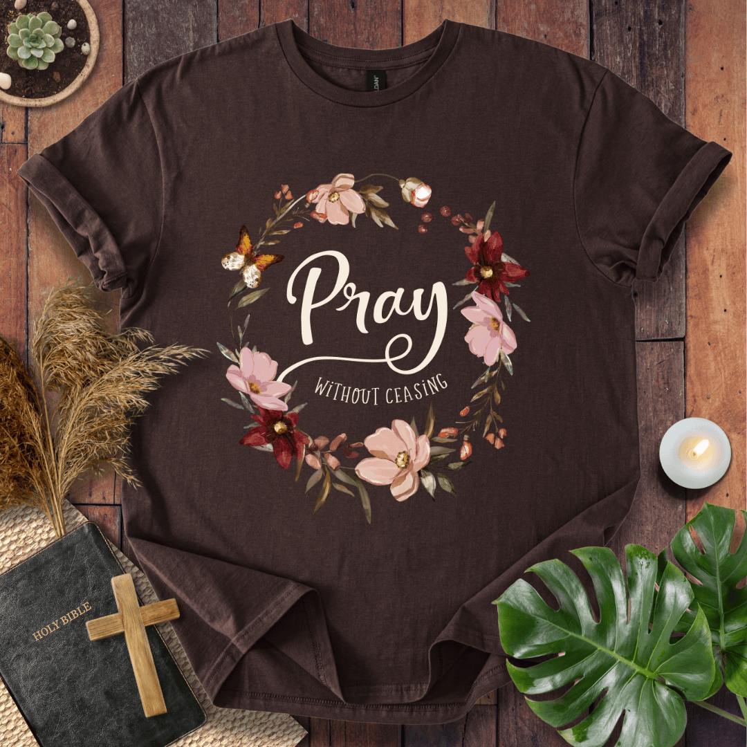 "Pray Without Ceasing" text inside a floral wreath with a butterfly on a shirt next to a Bible and cross.