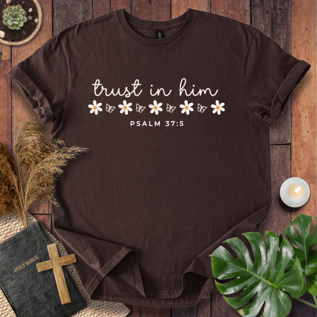 A dark brown short-sleeve t-shirt with a white graphic print reading 'trust in him' and 'PSALM 37:5' with small daisy illustrations, styled in a flat-lay on a wooden surface with a book, cross, candle, and green leaves.