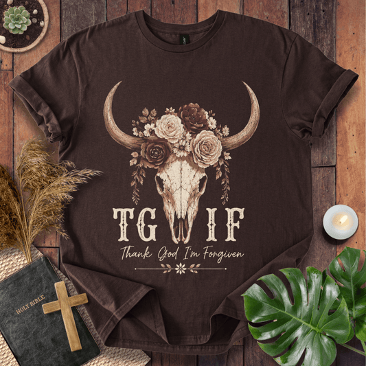A dark brown graphic t-shirt laid flat on a rustic wooden background, surrounded by decorative props including a Bible, a wooden cross, a lit candle, dried grass, and a green monstera leaf. The t-shirt features a graphic of a skull decorated with flowers and the text 'TG IF Thank God I'm Forgiven'.