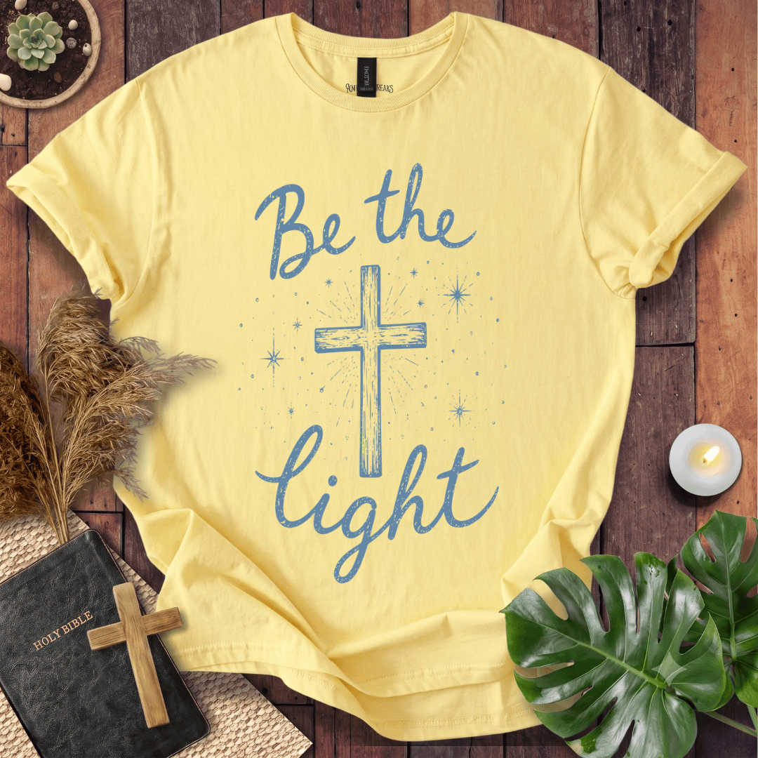 Christian t shirt for women with blue "Be the Light" text, a central cross, and starry accents on a wooden background.