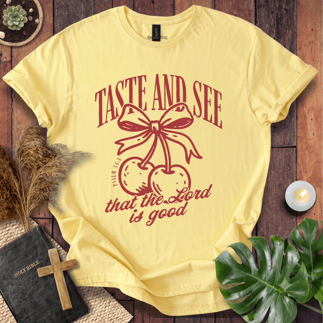 A pale yellow graphic T-shirt laid flat on a rustic wooden surface, featuring a red bow-and-cherry illustration with the text 'TASTE AND SEE that the Lord is good.' Decorative props including a book, cross, dried grass, candle, and green leaves surround the shirt.