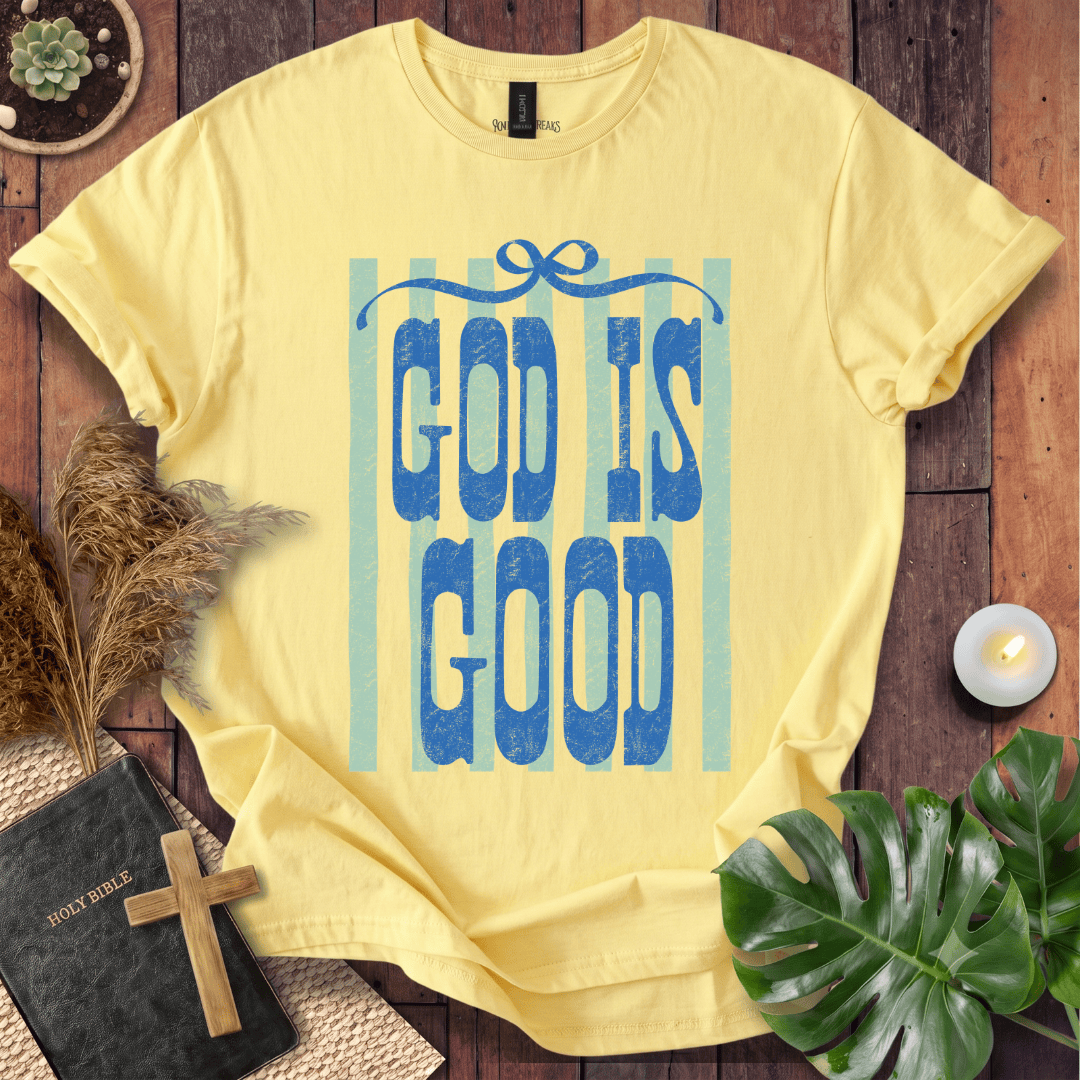 A pale yellow short-sleeve T-shirt laid flat on a rustic wooden surface, featuring a blue graphic that reads 'GOD IS GOOD' over alternating light blue and white vertical stripes. Decorative props around it include a small plant, dried grass, a candle, a black Bible with a wooden cross, and green leaves.