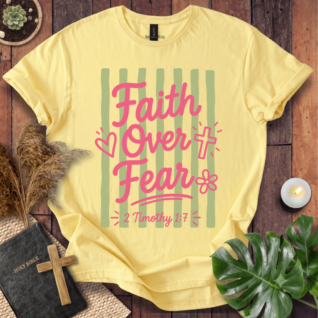 A pale yellow graphic T-shirt laid flat on a rustic wood surface, surrounded by decorative props including a Bible with a cross, dried grass, a candle, and green leaves. The shirt has a pink and green striped design with the words 'Faith Over Fear' and '2 Timothy 1:7'.