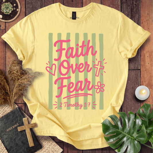 A pale yellow graphic T-shirt laid flat on a rustic wood surface, surrounded by decorative props including a Bible with a cross, dried grass, a candle, and green leaves. The shirt has a pink and green striped design with the words 'Faith Over Fear' and '2 Timothy 1:7'.