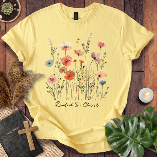 A pale yellow short-sleeve T-shirt laid flat on a rustic wooden surface, featuring a watercolor-style floral print with pink, orange, blue, and white flowers and the words 'Rooted In Christ' beneath them. Decorative props including a book with a wooden cross, pampas grass, a lit candle, and green leaves surround the shirt.