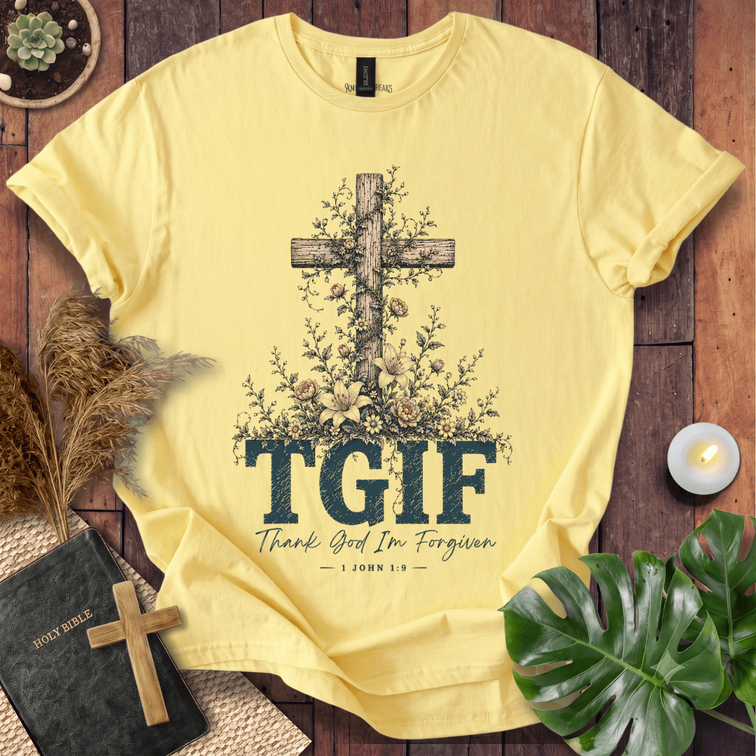 A pale yellow graphic T-shirt laid flat on a rustic wood surface, surrounded by decorative props including a Bible, candle, pampas grass, leaves, and a small cross. The shirt features a large wooden cross with flowers and the text 'TGIF' and 'Thank God I'm Forgiven — JOHN 1:9'.