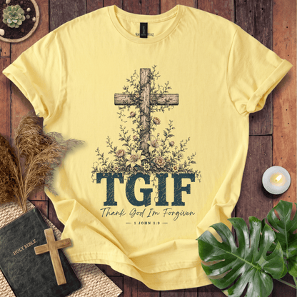 A pale yellow graphic T-shirt laid flat on a rustic wood surface, surrounded by decorative props including a Bible, candle, pampas grass, leaves, and a small cross. The shirt features a large wooden cross with flowers and the text 'TGIF' and 'Thank God I'm Forgiven — JOHN 1:9'.