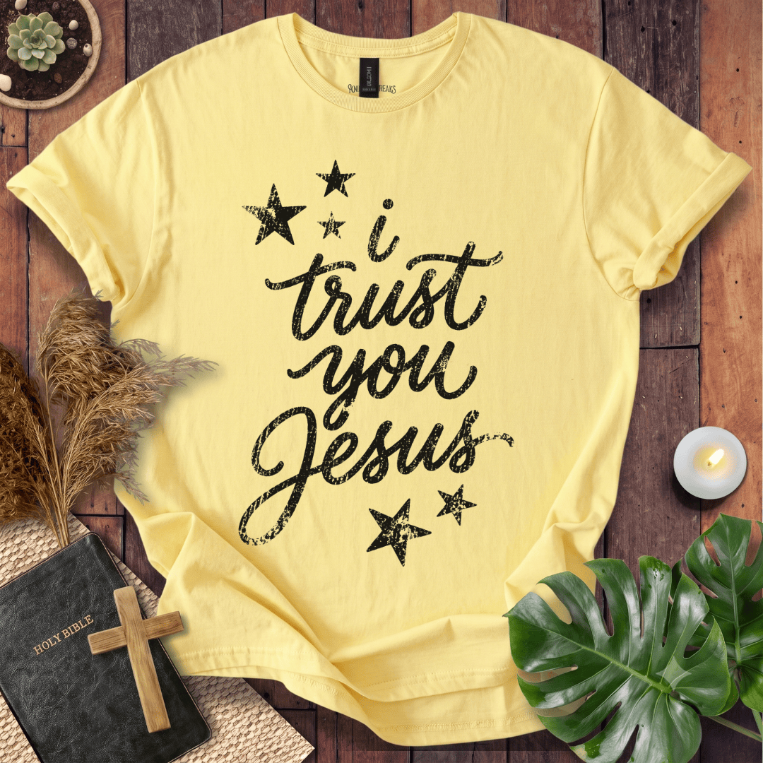 A pale yellow short-sleeve T-shirt laid flat on a rustic wooden surface, printed with the phrase 'I trust you Jesus' in black script and surrounded by black stars. The shirt is styled with a Bible, cross, pampas grass, a monstera leaf, and a small candle.