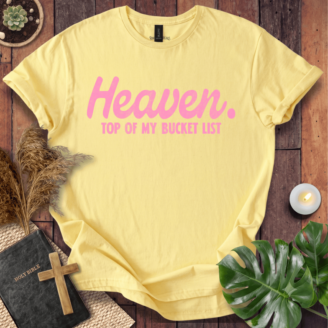 A pale yellow short-sleeve T-shirt laid flat on a rustic wooden surface, printed with the words 'Heaven. TOP OF MY BUCKET LIST' in pink.